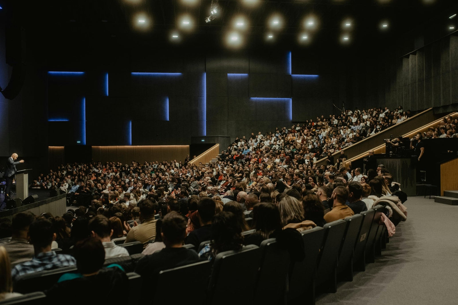 Large auditorium filled with spectators facing a stage with a speaker.