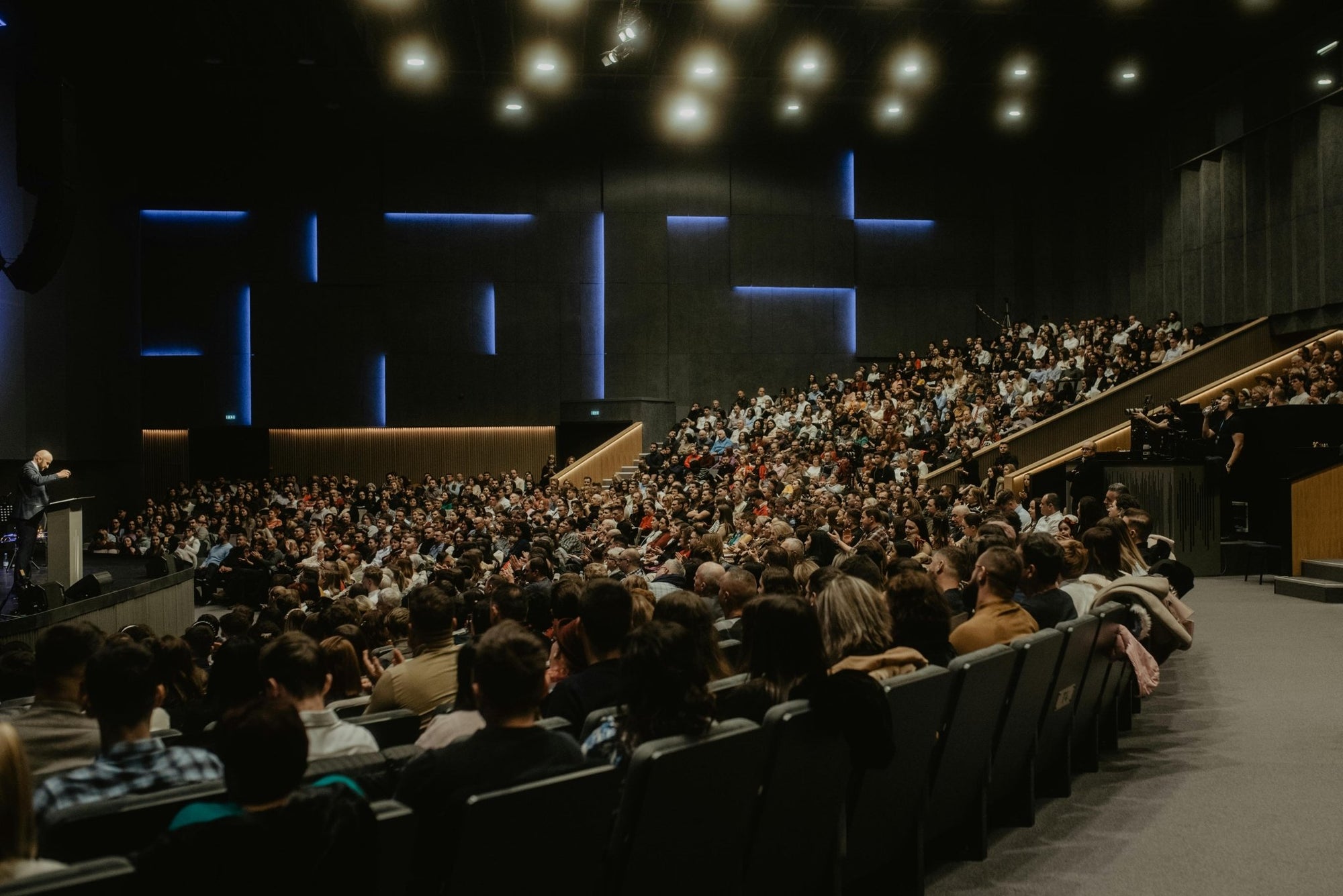 Large auditorium filled with spectators facing a stage with a speaker.