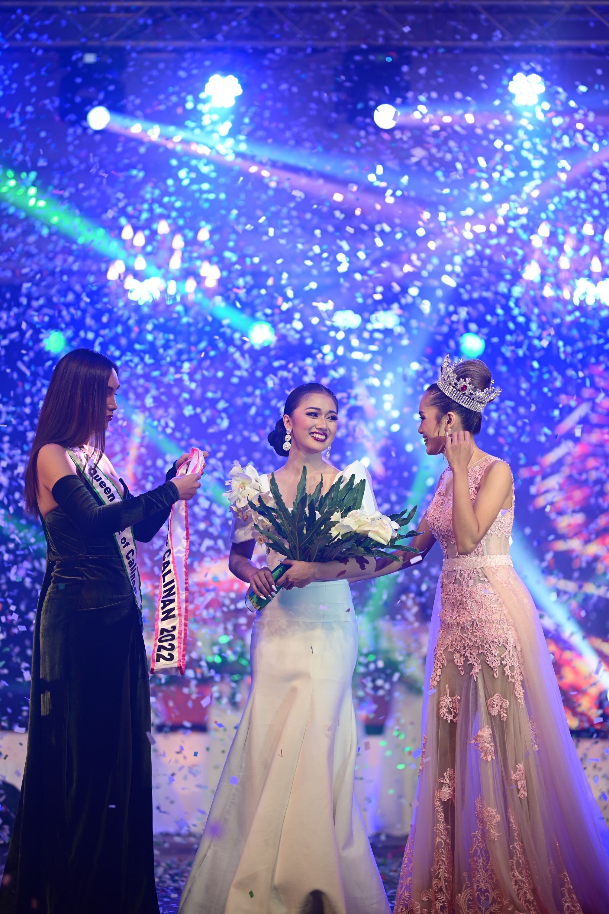 Three women on a brightly lit stage under blue and purple spotlights with confetti falling. The central woman wears a white gown and holds a bouquet, flanked by two other women in formal dresses, one wearing a sash.