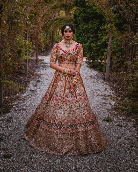 A woman stands on a gravel path surrounded by a leafy tunnel, wearing an ornate bronze and gold embroidered traditional lehenga choli set with pink and green floral detailing.