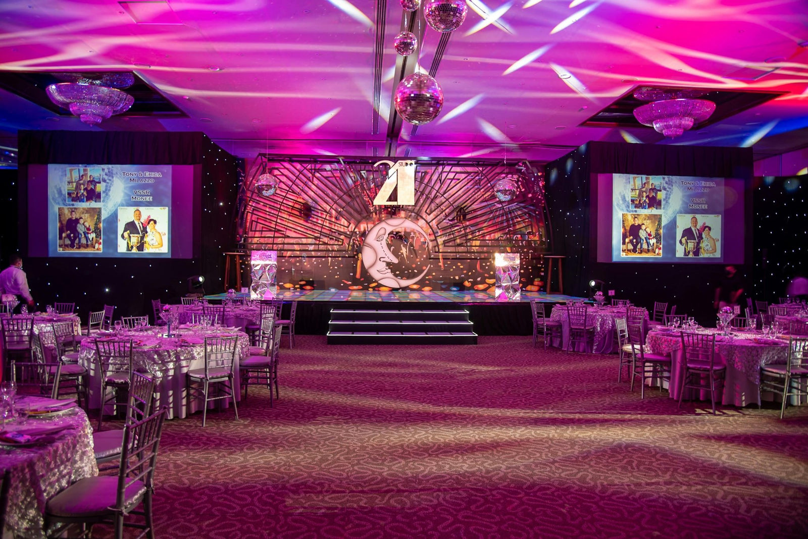 Elegant banquet hall with purple lighting, decorated tables, stage and disco balls.