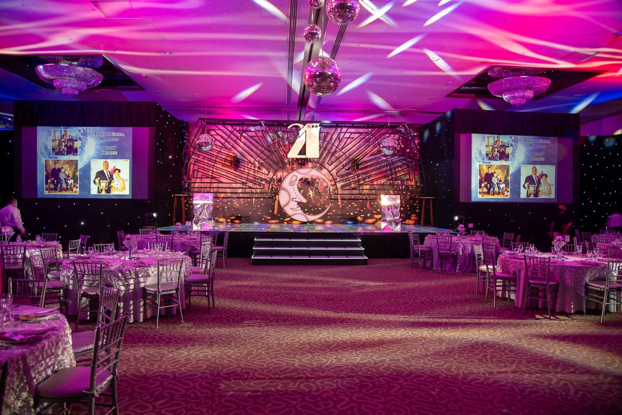 Elegant banquet hall with purple lighting, decorated tables, stage and disco balls.