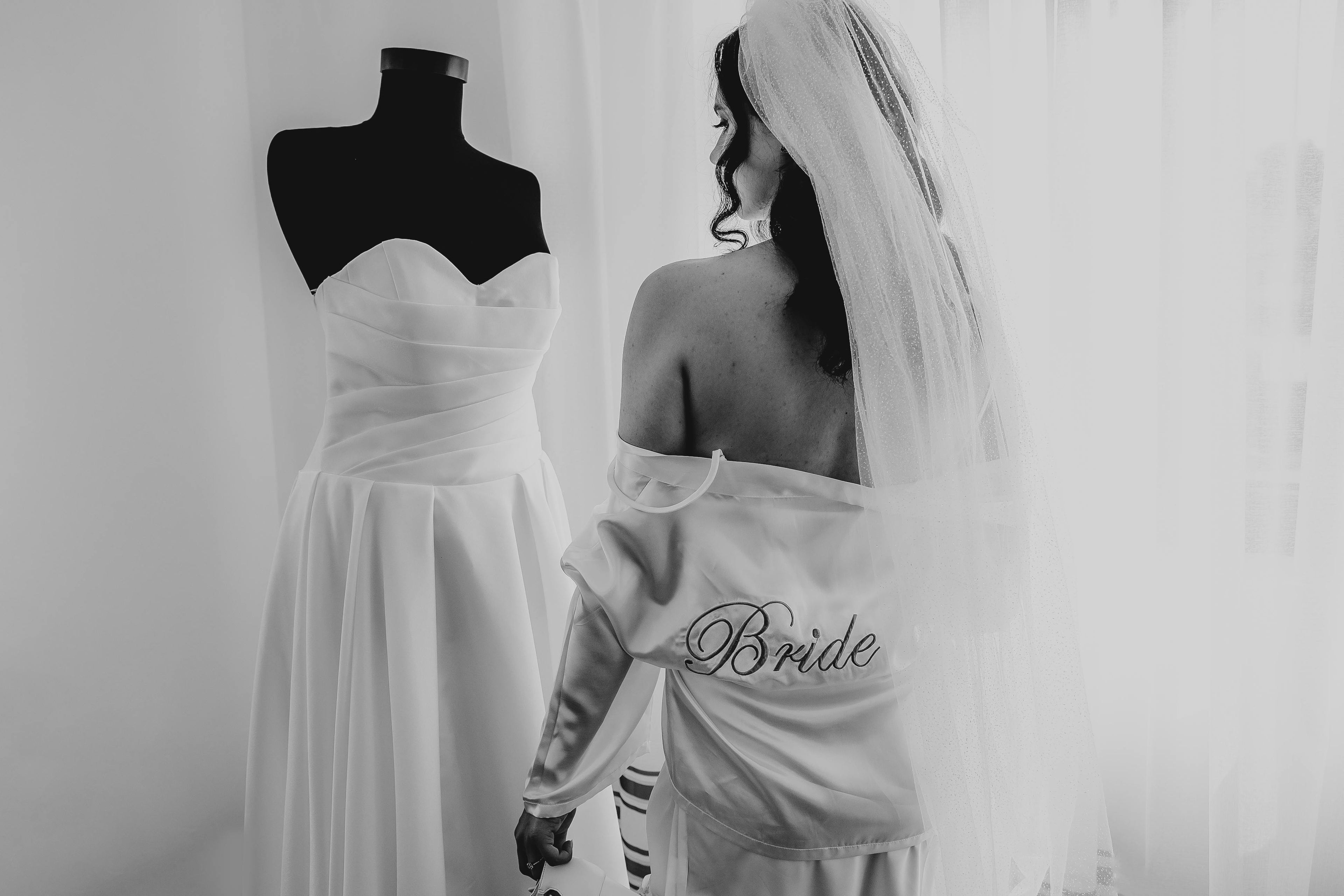 Woman wearing a 'Bride' robe with a white dress on a mannequin in the background.